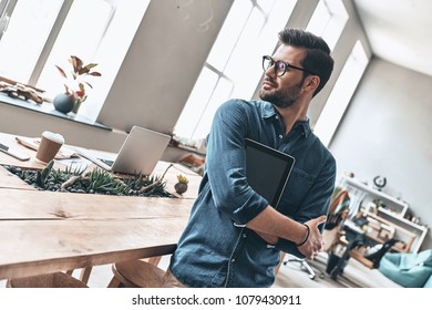 Thinking About Solution. Handsome Young Man Holding Digital Tablet And Looking Away With Smile While Standing In The Office       