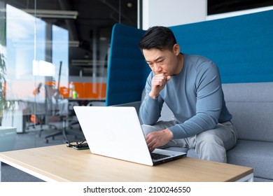 A Thinking Asian Man Works At A Computer In A Modern Office, A Freelancer Is Focused On Solving A Task, Makes An Important Job.