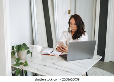 Thinking Brunette Woman With Long Hair In White Cardigan Working On Laptop Using Mobile Phone In Bright Modern Office