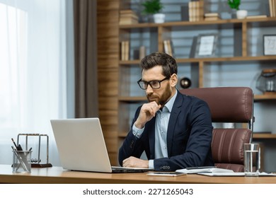 Thinking businessman working sitting at desk, mature adult boss in business suit and beard looking at laptop screen thinking about financial investment decisions inside office. - Shutterstock ID 2271265043