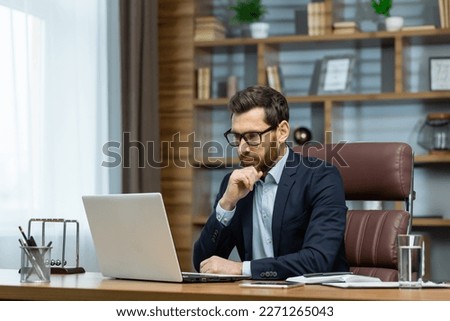 Thinking businessman working sitting at desk, mature adult boss in business suit and beard looking at laptop screen thinking about financial investment decisions inside office.