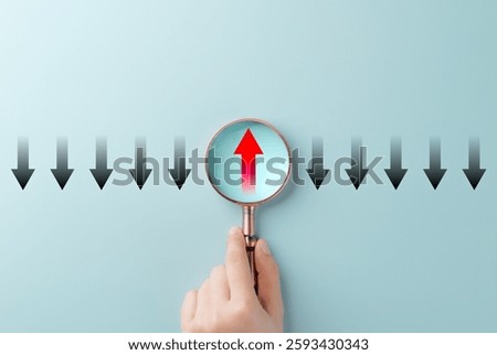 Thinking differently and finding new perspectives. A Hand holding magnifying glass reveals a red upward arrow among black downward arrows, innovation, leadership, unique thinking, and problem-solving.