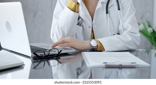 Thinking doctor woman is reviewing medical records on laptop. Close-up of a healthcare unknown professional analyzing patient information on a computer. Medicine - Powered by Shutterstock