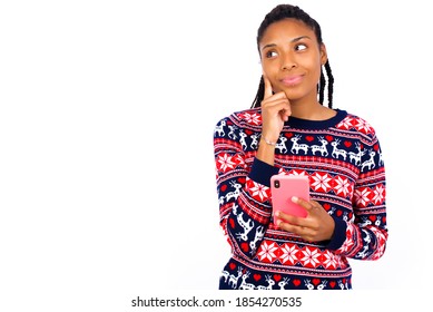 Thinking Dreaming Excited Young Beautiful African American Woman Wearing Christmas Sweater, Against White Wall Using Mobile Phone And Holding Hand On Face. Taking Decisions And Social Media Concept.