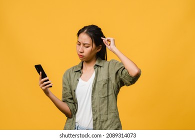 Thinking Dreaming Young Asia Lady Using Phone With Positive Expression, Dressed In Casual Cloth Feeling Happiness And Stand Isolated On Yellow Background. Happy Adorable Glad Woman Rejoices Success.