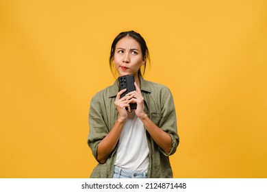 Thinking Dreaming Young Asia Lady Using Phone With Positive Expression, Dressed In Casual Cloth Feeling Happiness And Stand Isolated On Yellow Background. Happy Adorable Glad Woman Rejoices Success.