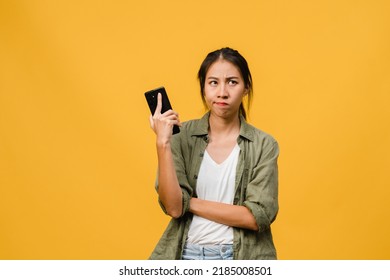 Thinking dreaming young Asia lady using phone with positive expression, dressed in casual cloth feeling happiness and stand isolated on yellow background. Happy adorable glad woman rejoices success. - Shutterstock ID 2185008501