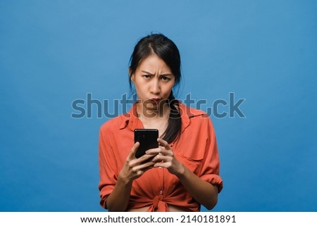 Thinking dreaming young Asia lady using phone with positive expression, dressed in casual clothing feeling happiness and stand isolated on blue background. Happy adorable glad woman rejoices success.