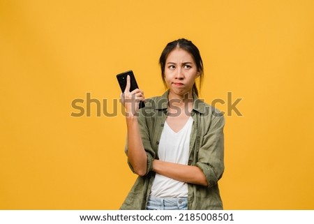 Thinking dreaming young Asia lady using phone with positive expression, dressed in casual cloth feeling happiness and stand isolated on yellow background. Happy adorable glad woman rejoices success.