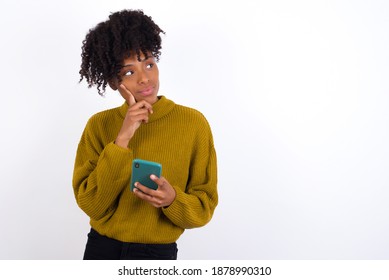 Thinking dreaming Young beautiful African American woman wearing knitted sweater against white wall using mobile phone and holding hand on face. Taking decisions and social media concept. - Shutterstock ID 1878990310