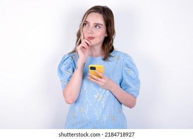 Thinking dreaming young caucasian woman wearing blue T-shirt over white background using mobile phone and holding hand on face. Taking decisions and social media concept.