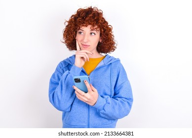 Thinking dreaming young redhead girl wearing blue jacket over white background  using mobile phone and holding hand on face. Taking decisions and social media concept. - Shutterstock ID 2133664381