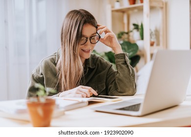 Thinking Freelance Woman In Glasses With Phone And Note Book Typing At Laptop And Working From Home Office With Plants. Happy Girl On Workplace At The Desk. Distance Learning Online Education And.