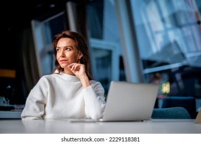 Thinking middle aged business woman using laptop while sitting at desk at the office. Executive businesswoman wearing sweater.  - Powered by Shutterstock