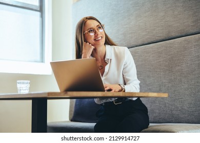 Thinking of new exciting business ideas. Happy young businesswoman smiling cheerfully while looking away. Young female entrepreneur working on a laptop in a modern workspace. - Powered by Shutterstock