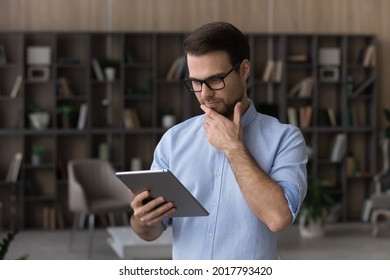 Thinking on information. Concentrated millennial male in eyewear engaged in reading online book on digital tablet pc using e reader app. Pensive young man ponder on business news on corporate website - Powered by Shutterstock