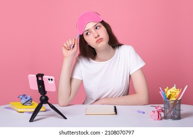 thinking thoughtful brunette teen school girl doing homework scratches head with pencil sit desk use smartphone and phone stand for online lessons wearing pink sleep mask distance education concept
