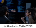 Thinking through a technical problem during a late night meeting, two developers face monitors with source code and charts, lit by soft blue and warm desk lamp light.