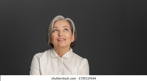 Thinking White-haired Business Woman Looking Upward. Creative Middle-aged Pretty Woman Thoughtfully Smiles Posing On Grey Background. Close Up Portrait. Copyspace At Right Side.