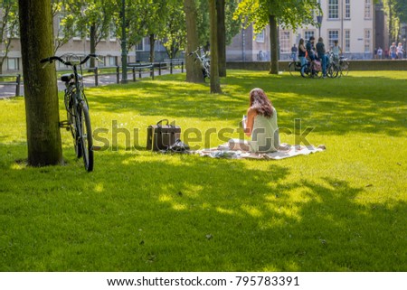 Thinly dressed unknown young woman sitting on a plaid in the grass looks in the Dutch park on her smartphone. Her bike is parked against a tree. In the background a few teenagers are talking.