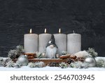 Third Advent: Decoration with four grey Advent candles, a gnome and Christmas decorations in the snow in front of a dark slate wall with space for text.