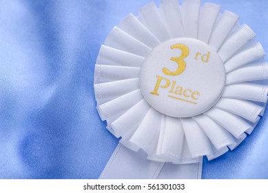 Third place winners white rosette with gold text and a pleated ribbon in a close up oblique angle view conceptual of competition and achievement