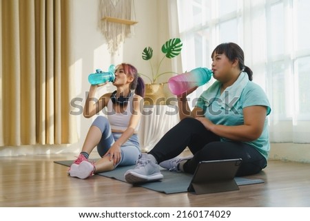 Thirst while exercising concept. Two Asian women's body size is different in sportswear, drinking water while resting and sitting on yoga mat after fitness exercise at home together.