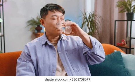 Thirsty asian man holding glass of natural aqua make sips drinking still water preventing dehydration sits at home living room. Chinese Guy with good life habits, healthy slimming, weight loss concept