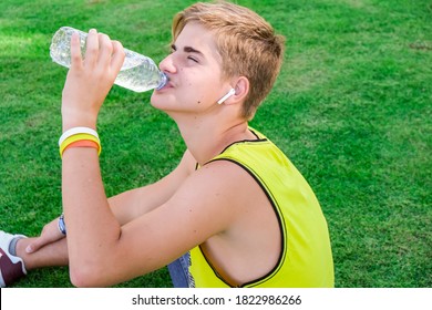 Thirsty Athlete Drinking Water After Workout. Young Man, Teen Drink Clear Blue Water From A Plastic Bottle After A Workout Sitting On A Green Grass, Refreshing, Relax. Sport, Healthy Life Style.