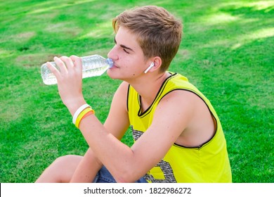 Thirsty Athlete Drinking Water After Workout. Young Man, Teen Drink Clear Blue Water From A Plastic Bottle After A Workout Sitting On A Green Grass, Refreshing, Relax. Sport, Healthy Life Style.