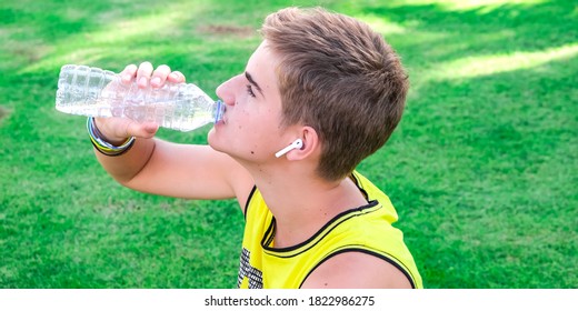 Thirsty Athlete Drinking Water After Workout. Young Man, Teen Drink Clear Blue Water From A Plastic Bottle After A Workout Sitting On A Green Grass, Refreshing, Relax. Sport, Healthy Life Style.