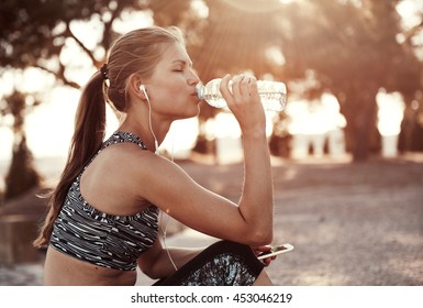 Dreißige Joggerin trinkt frisches Wasser und hört nach dem Training Musik in Kopfhörern. Junge Sportlerin, die im Park draußen trainiert. 