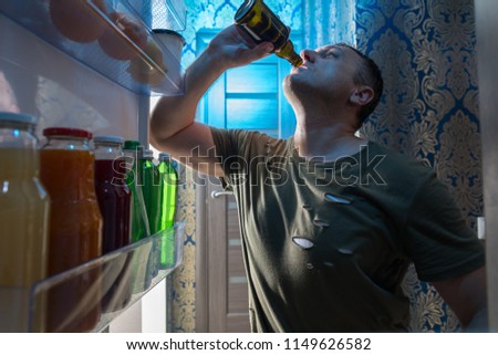 Thirsty man gulping down a cold beer as he stands in the open door of his fridge viewed from inside looking out