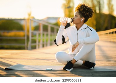 Thirsty Young African American Woman Drinking Water During Outdoor Training, Sporty Black Female Sitting On Fitness Mat And Enjoying Healthy Drink, Resting After Workout Outside, Copy Space - Powered by Shutterstock