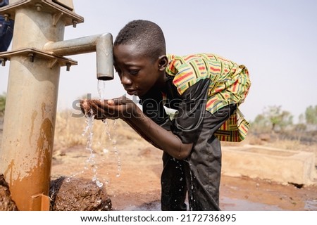 Thirsty young black African boy leans forward to drink fresh and clean water with his bare hands from a tap; water scarcity concept