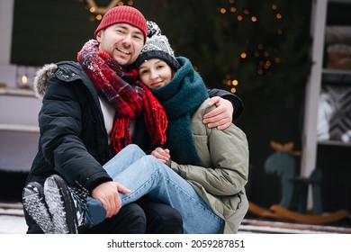 Thirty Year Old Couple Sitting On A Snowy Terrace Step In Front Of The House, Posing For A Photo. Both Wearing Hats, Jackets And Scarfs. He's Holding Her Ankles On His Knees.