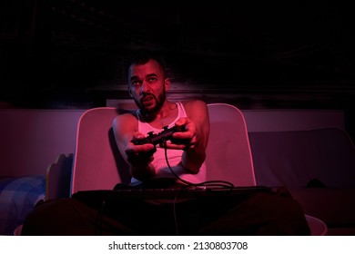 A Thirty-year-old Guy With A Joystick In His Hands Is Playing Computer Games, Experiencing Various Emotions. Sitting On The Couch At Home. View From The TV Side.