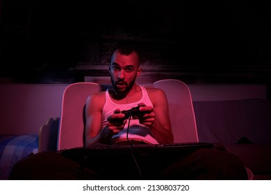 A Thirty-year-old Guy With A Joystick In His Hands Is Playing Computer Games, Experiencing Various Emotions. Sitting On The Couch At Home. View From The TV Side.