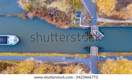 This aerial image captures a boat floating close to a small, simple river dock. The waterway appears calm and is bordered by autumnal vegetation. A road intersects the frame, leading to the dock and