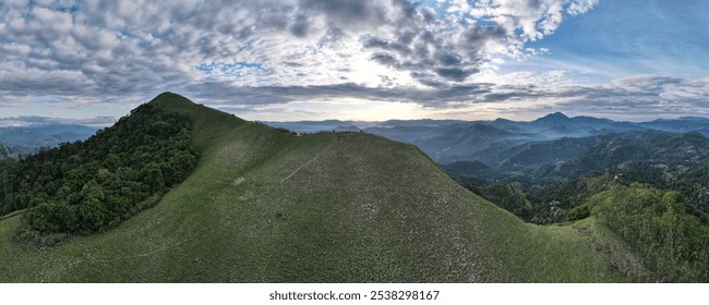 This aerial image captures a lush green mountain range with rolling hills, dense forests, and misty layers of distant peaks under a partly cloudy sky.  - Powered by Shutterstock