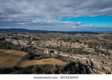 This aerial image captures a panoramic view of a densely built historic town nestled amidst rolling hills and green countryside in Italy. The scene features traditional architecture, agricultural fields, and distant mountains under a dynamic sky with both blue patches and fluffy clouds.