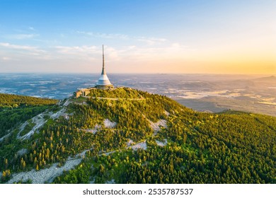 This aerial view captures Jested mountain in Liberec, Czechia, showcasing the iconic hotel and TV transmitter amid sprawling greenery at sunset, revealing stunning landscapes.