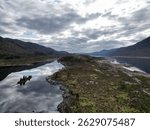 This aerial view captures the serene beauty of Loch Cluanie in Scotland. A rugged landmass, covered in low-lying vegetation and dotted with rocky outcrops, extends into the still, dark water. 