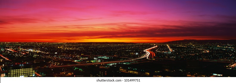 This Is An Aerial View Of Downtown Los Angeles At Sunset. The Streaked Lights Of The Freeway Are In The Center With An Orange Sunset Sky.