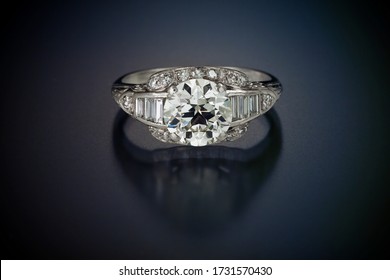 This antique diamond engagement ring with a one plus carat center stone is enhanced by accent side diamonds and set in platinum. Shown laying on a black reflective background.