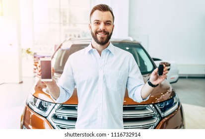 With This App On Mobile Much Easier Buying A Car. Smiling Handsome Customer In Dealership Shows Smart Phone And Car Keys In Camera