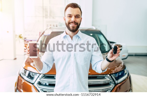 With this app on mobile much easier buying a car.\
Smiling handsome customer in dealership shows smart phone and car\
keys in camera