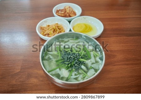 This appetizing culinary imagery captures authentic Daseulgi Sujebi a traditional hand torn dough soup from Jangsu county in South Korea. The stainless bowl presents a clear nutritious broth filled with chewy wheat flakes fresh green chives and nutrient rich freshwater marsh snails. A close up macro shot emphasizes a metal spoon lifting the delicate noodles highlighting its comforting texture. Accompanied by classic banchan side dishes like kimchi and pickled radish on a rustic wooden table this healthy local gastronomy is perfect for Asian food editorials and regional restaurant menus.