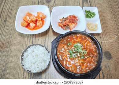 This appetizing culinary visual captures a hearty serving of spicy beef tripe soup known as naejang gukbap in South Korea Served in a traditional black earthen vessel on a rustic wooden table the steaming red broth is packed with tender offal enoki mushrooms and green scallions A macro shot highlights a metal spoon lifting savory rice mixed with the flavorful stew and chewy meat textures Accompanied by classic banchan like cubed radish kimchi and chopped chilies this comforting regional meal is ideal for Asian gastronomy projects local restaurant menus and food editorials worldwide
