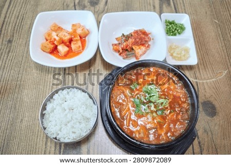 This appetizing culinary visual captures a hearty serving of spicy beef tripe soup known as naejang gukbap in South Korea Served in a traditional black earthen vessel on a rustic wooden table the steaming red broth is packed with tender offal enoki mushrooms and green scallions A macro shot highlights a metal spoon lifting savory rice mixed with the flavorful stew and chewy meat textures Accompanied by classic banchan like cubed radish kimchi and chopped chilies this comforting regional meal is ideal for Asian gastronomy projects local restaurant menus and food editorials worldwide
